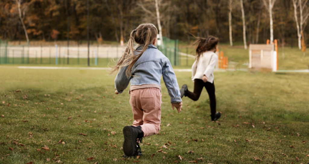 Zwei Kinder rennen von hinten gesehen über eine weite grüne Wiese in Richtung eines herbstlichen Waldes.