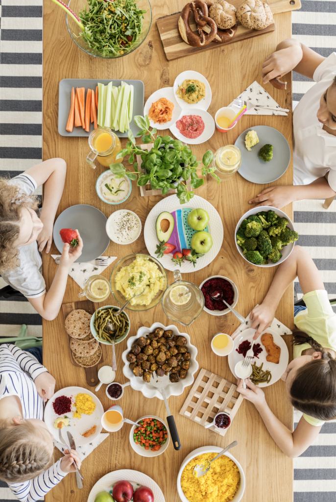 Vogelperspektive eines Holztisches mit gesundem Essen wie Brokkoli, Avocado, Äpfeln und Dips, um den drei Kinder sitzen.