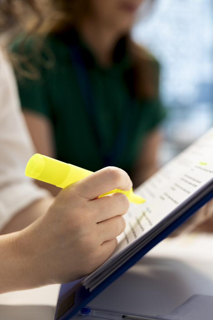 Nahaufnahme einer Hand, die mit einem gelben Textmarker eine Zeile in einem Buch oder Dokument auf einem Klemmbrett markiert.