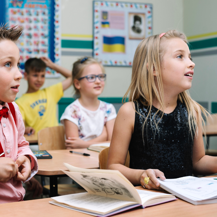 Mehrere Kinder sitzen an ihren Schreibtischen in einem hellen Klassenzimmer. Ein Mädchen mit blonden Haaren im Vordergrund blickt aufmerksam nach oben, während sie ein offenes Buch vor sich hat. Im Hintergrund sind weitere Kinder und pädagogische Poster an der Wand zu sehen.